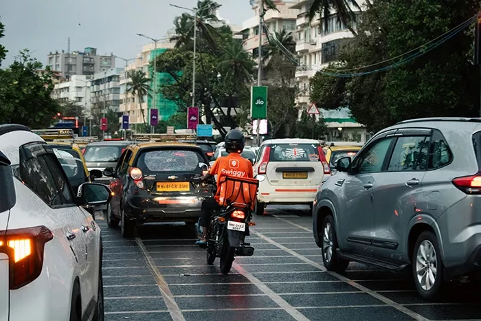 A Swiggy delivery rider in a bright orange t-shirt navigates through dense South Mumbai traffic on his motorbike.