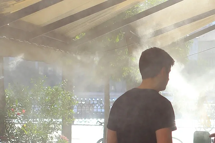 A man under a cafe awning is enveloped in mist from a cooling sprinkler
