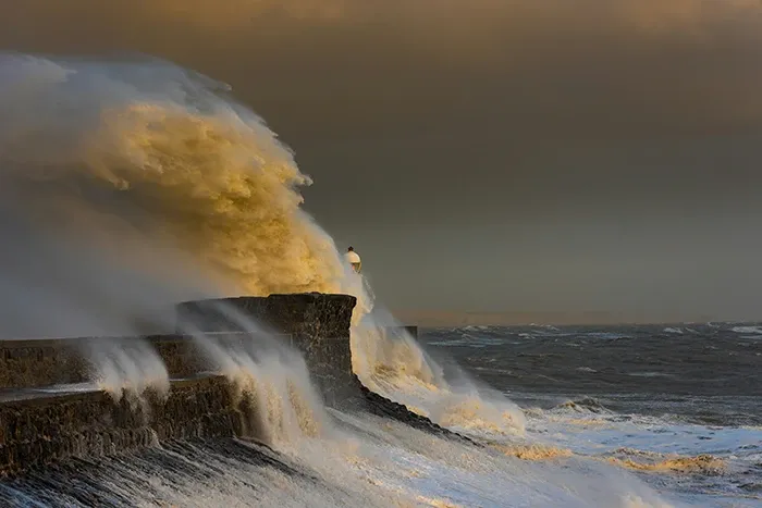 A large wave strikes the shore near a lighthouse in Wales