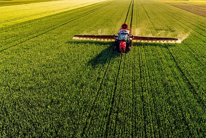 Drone shot of a red tractor applying treatment to wheat crops in vast farmland. oticki/AdobeStock.
